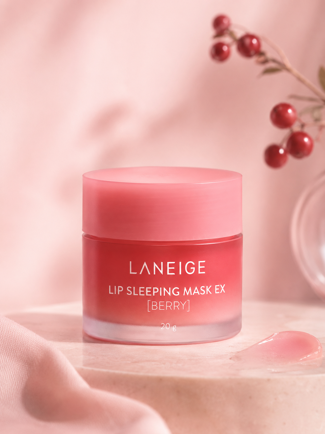 A LANEIGE Lip Sleeping Mask EX Berry jar sits on a marble surface, surrounded by soft fabric and decorative berries, showcasing its vibrant pink color and elegant packaging.