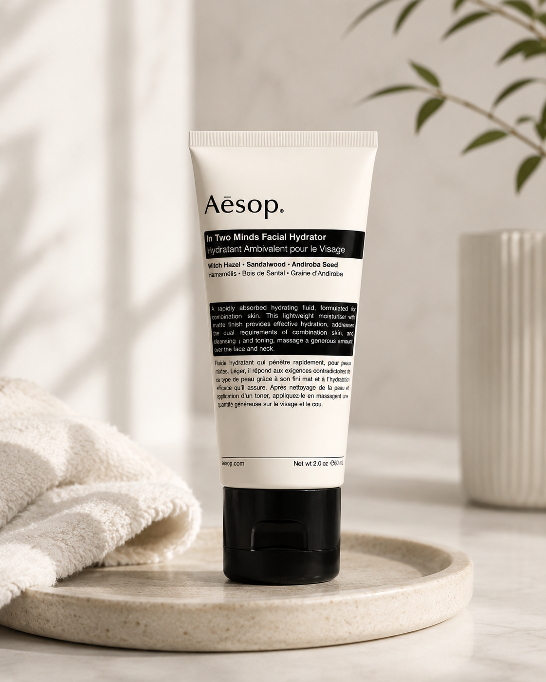Aesop In Two Minds Facial Hydrator displayed on a minimalist stone tray in a bright, spa-inspired bathroom setting with soft natural light, a folded white towel, ceramic accessories, and botanical greenery. The clean, calming composition highlights the lightweight moisturizer as part of a modern skincare routine, emphasizing balanced hydration and a fresh matte finish for combination skin.