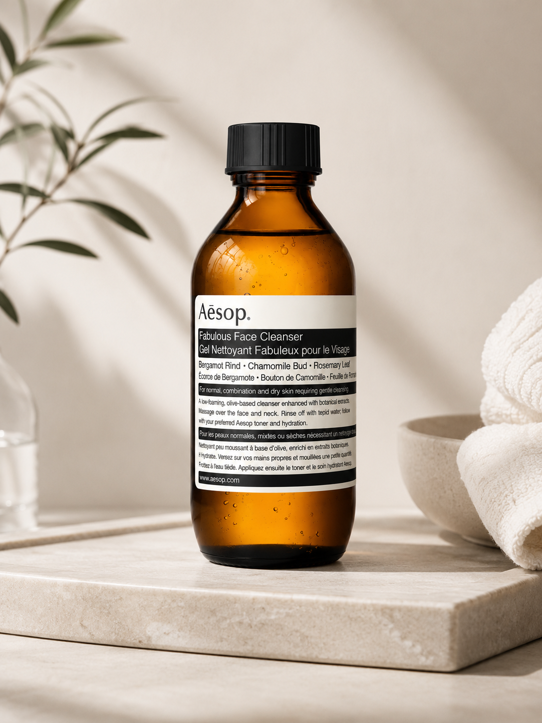 Aesop Fabulous Face Cleanser displayed on a natural stone surface in a calm, minimalist skincare setting with soft natural light, ceramic accessories, folded white towels, and botanical greenery. The warm ambient styling highlights the amber glass bottle and conveys the cleanser's gentle, botanical formulation for everyday facial cleansing.