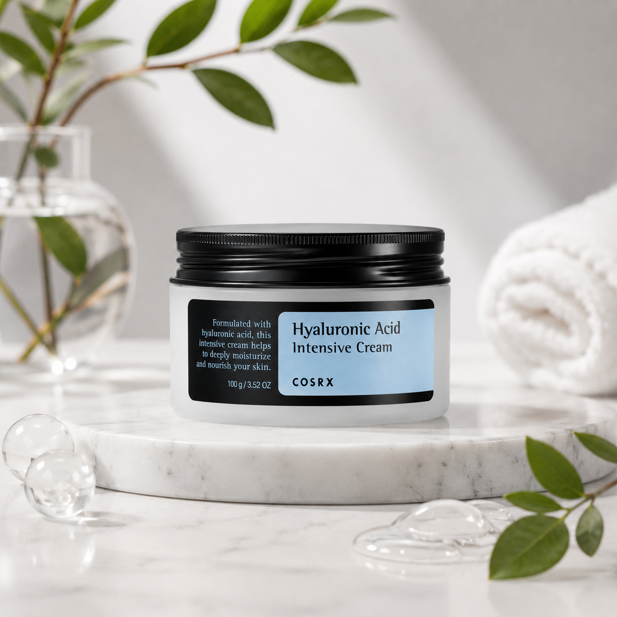 COSRX Hyaluronic Acid Intensive Cream presented on a marble surface with green botanical leaves, a glass vase, water droplets, and a rolled white towel, highlighting its hydrating skincare benefits. Enriched with hyaluronic acid, this intensive moisturizer helps replenish moisture, nourish dry skin, reinforce the skin barrier, and provide lasting hydration for a soft, healthy-looking, and radiant complexion while maintaining a lightweight, comfortable feel.