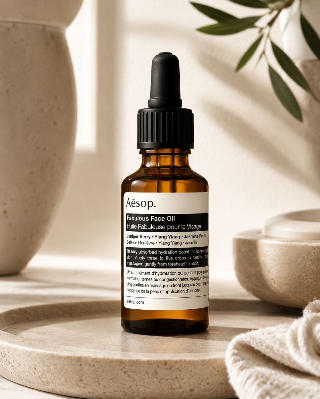 Aesop Fabulous Face Oil displayed on a natural stone tray in a softly lit minimalist skincare setting, surrounded by ceramic vessels, botanical olive branches, and neutral textures. The warm ambient lighting highlights the amber glass bottle and emphasizes the product's luxurious botanical formulation for nourishing and hydrating the skin.
